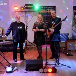 Trzyosobowy zespół muzyczny w trakcie występu: mężczyzna grający na gitarze elektrycznej, kobieta z mikrofonem i mężczyzna grający na keyboardzie Korg, oświetlenie sceniczne i baner reklamowy...