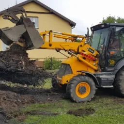 Żółta koparko-ładowarka JCB 3CX podnosi korzeń drzewa na tle domu i zieleni.