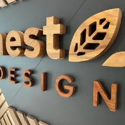 Drewniane litery tworzące napis 'nest DESIGN' na szarej ścianie, z elementem dekoracyjnym w kształcie liścia z wyciętymi wzorami, całość wykonana z różnych odcieni drewna.