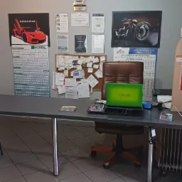 Biuro warsztatu samochodowego z biurkiem, laptopem Sony Vaio, krzesłem obrotowym i ścianą z dyplomami, certyfikatami oraz plakatem z czerwonym samochodem i motocyklem.