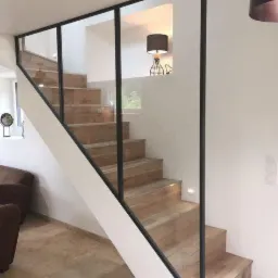 Nowoczesne schody drewniane z przeszkloną balustradą o czarnych profilach; widoczne oświetlenie schodów i lampa w tle.