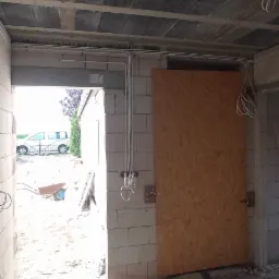 Wnętrze surowego budynku w trakcie prac, widoczne poprowadzone przewody elektryczne na ścianach i pod sufitem, ściany z białego bloczka, podłoga w stanie surowym.