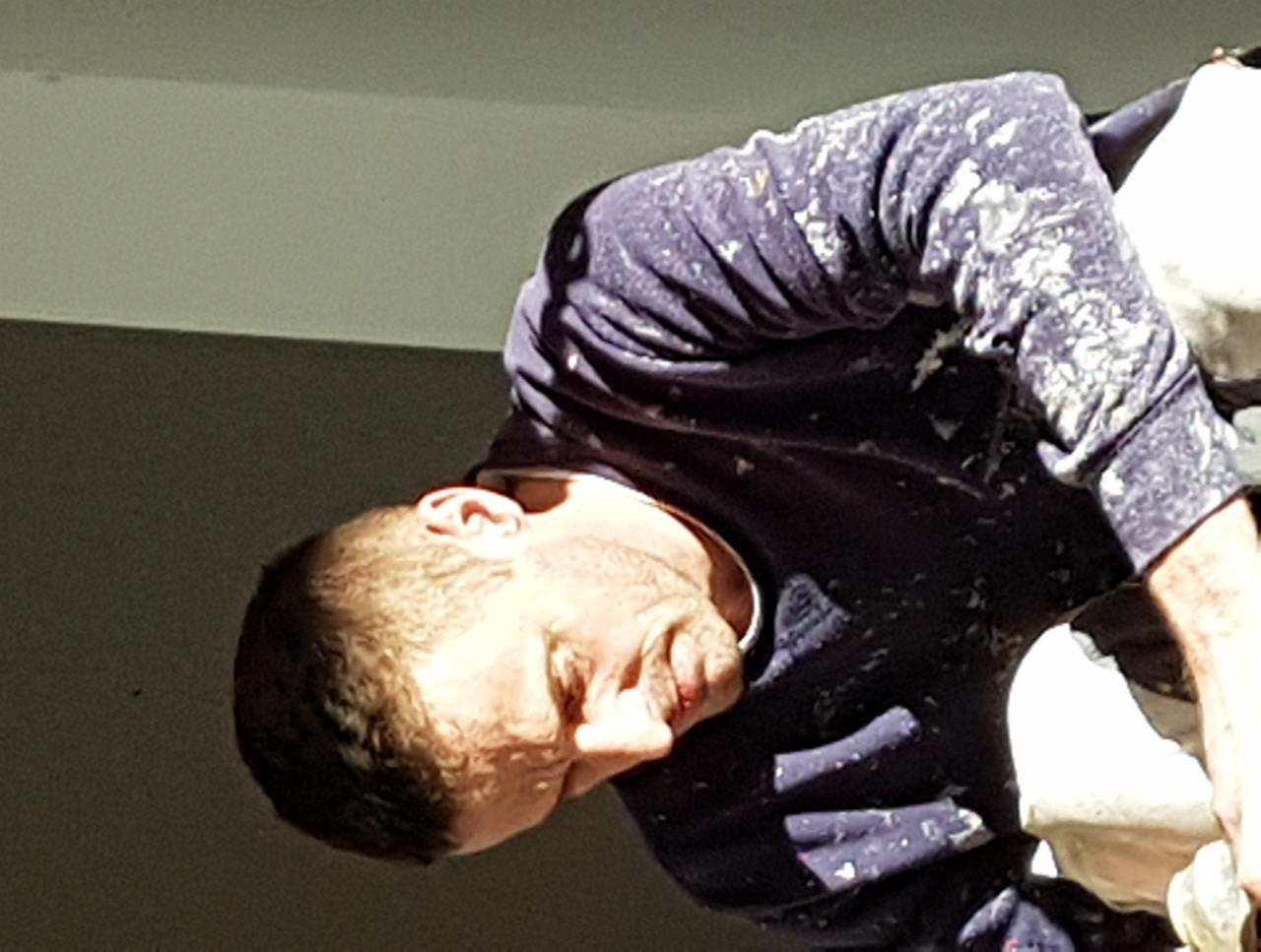 Mężczyzna w bluzie ubrudzonej farbą, pochylony, prawdopodobnie podczas malowania wnętrza, widoczny fragment ściany i sufitu w tle.