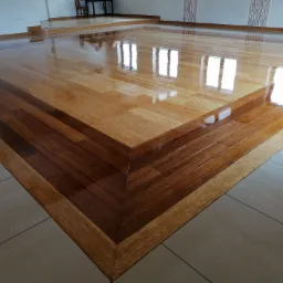 Wypolerowana drewniana podłoga z wyraźnym wzorem ułożonych desek i odblaskami okien, otoczona ceramicznymi płytkami podłogowymi.