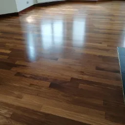 Błyszcząca, ułożona parkietowa podłoga z ciemnego drewna, z widocznymi refleksami światła padającego z okna, przy listwie przypodłogowej w kolorze drewna.