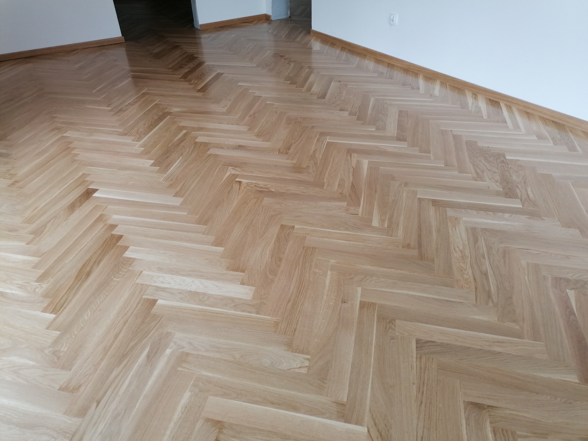 Jasny parkiet ułożony we wzór jodełki francuskiej, widoczne listwy przypodłogowe i fragment ściany.