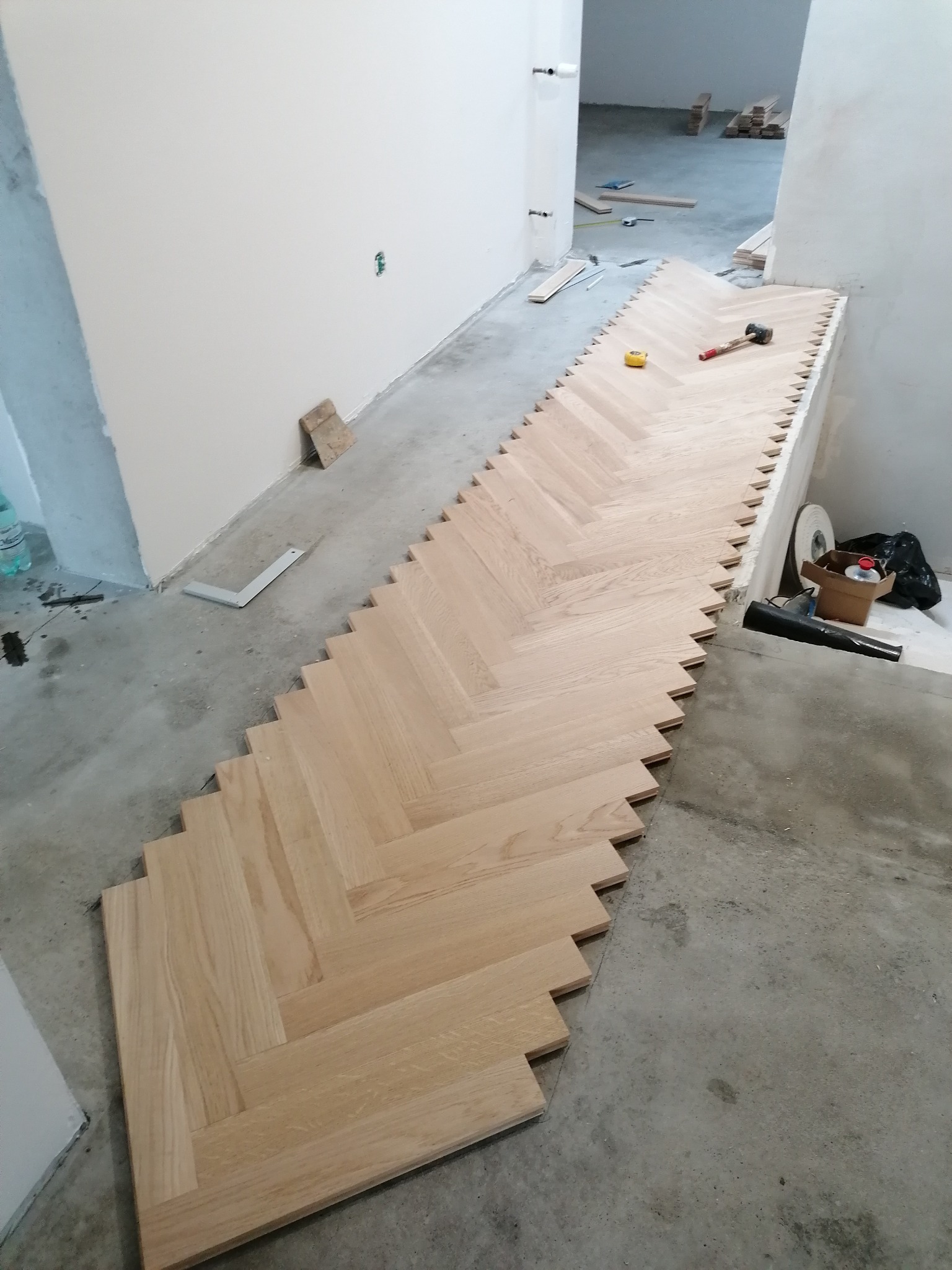 Fragment układanej drewnianej podłogi w jodełkę na surowym podłożu, widoczne narzędzia i materiały budowlane, etap prac wykończeniowych.