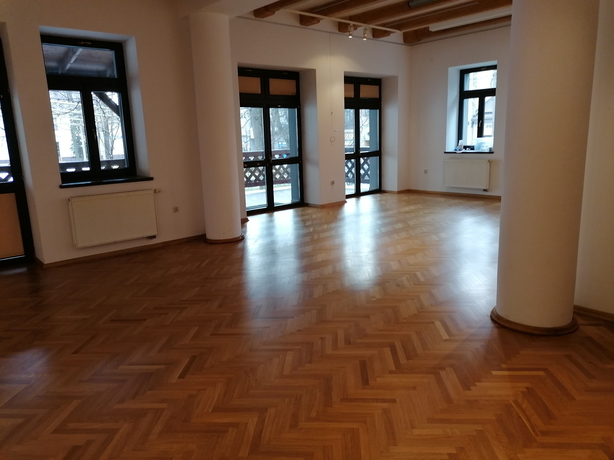 Parkiet ułożony we wzór jodełki w przestronnym, jasnym wnętrzu z kolumnami i dużymi oknami balkonowymi.