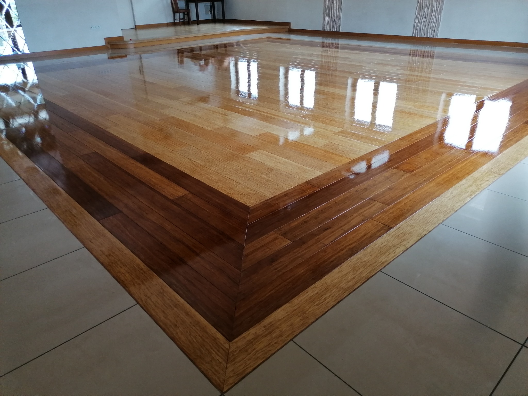 Wypolerowana drewniana podłoga z wyraźnym wzorem ułożonych desek i odblaskami okien, otoczona ceramicznymi płytkami podłogowymi.