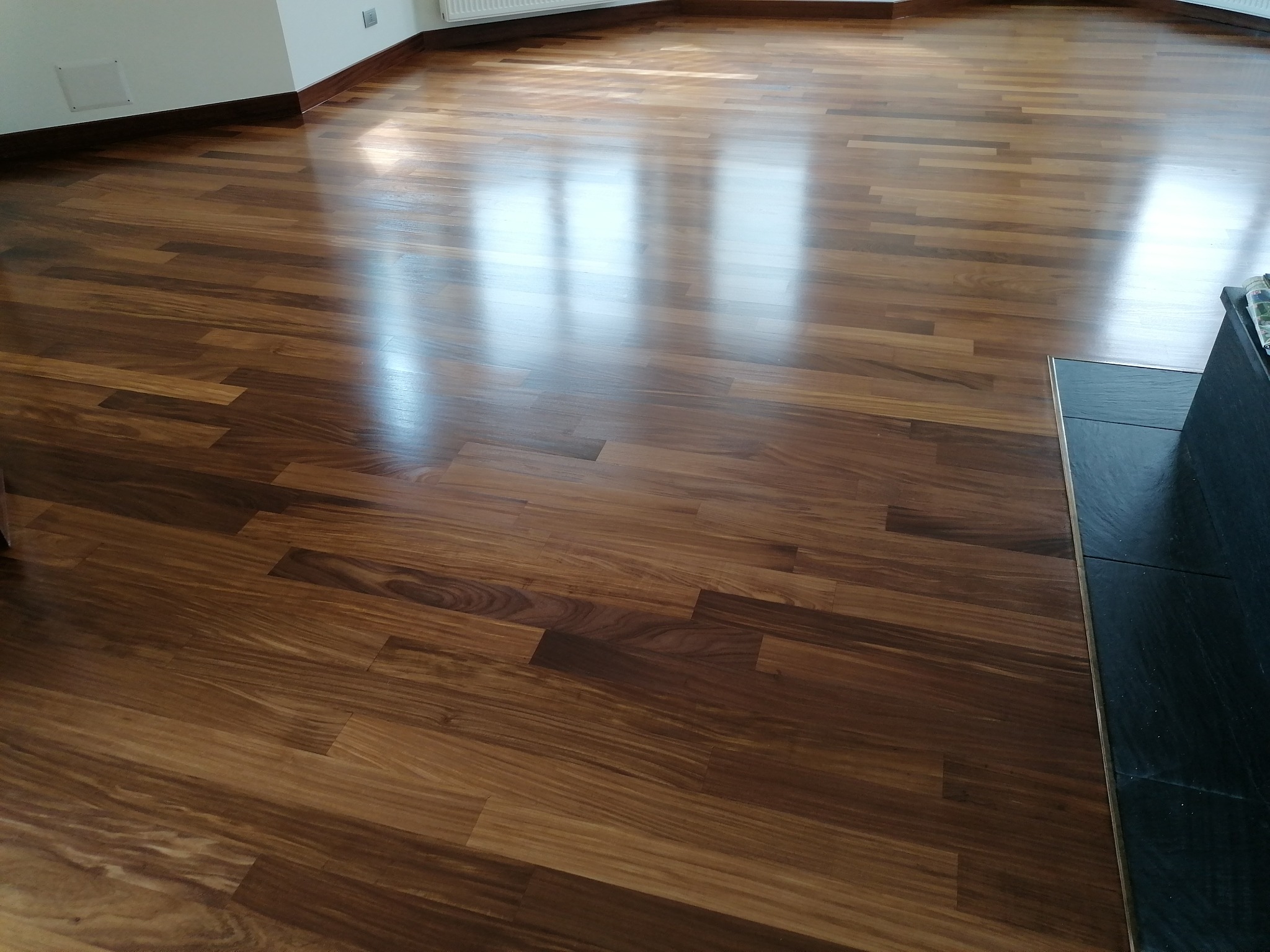 Błyszcząca, ułożona parkietowa podłoga z ciemnego drewna, z widocznymi refleksami światła padającego z okna, przy listwie przypodłogowej w kolorze drewna.