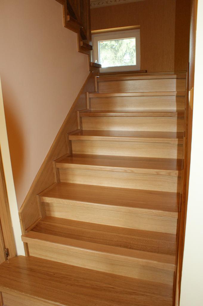 Jasne, drewniane schody z widocznym usłojeniem, prowadzące w górę, oświetlone przez okno w tle.