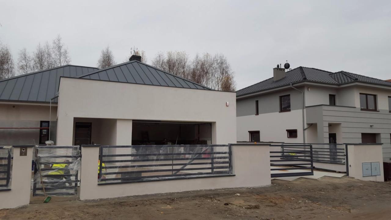 Dwa nowe domy jednorodzinne z szarymi dachami, ogrodzone metalowymi płotami na podmurówce; jeden dom z garażem w trakcie prac wykończeniowych, zabezpieczony folią.