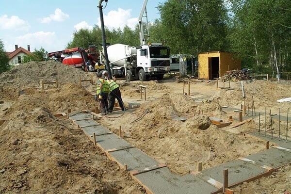 Wylewanie fundamentów pod budynek z użyciem betonomieszarki i pompy do betonu, widoczni pracownicy w kamizelkach odblaskowych, szalunki przygotowane pod wylewkę.