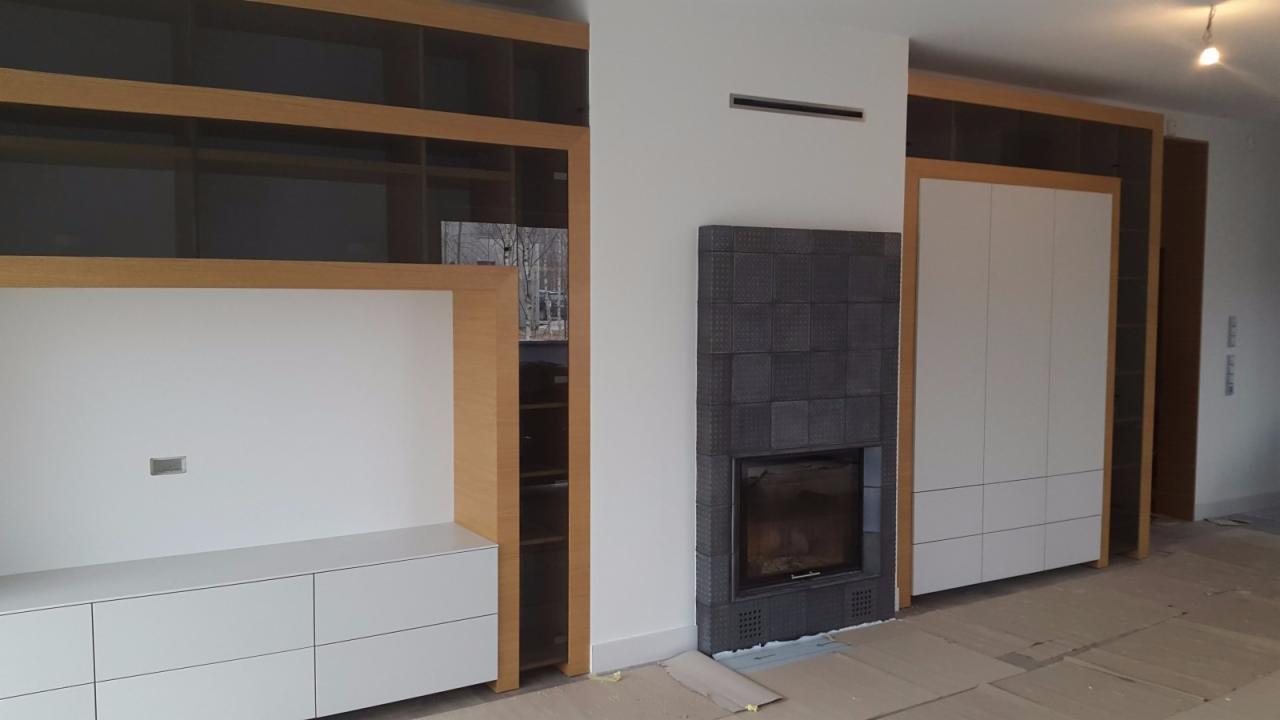 Nowoczesny salon z zabudową meblową z jasnego drewna i białymi frontami, kominek obłożony ciemną, kwadratową płytką, widoczne elementy wykończeniowe podłogi.