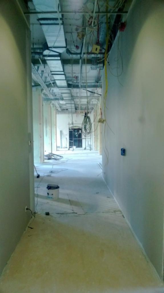 Korytarz biurowy w trakcie prac wykończeniowych: widoczne instalacje pod sufitem, surowe ściany, wiadro z farbą i luźne przewody.