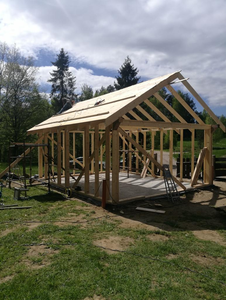 Szkielet drewnianej konstrukcji budynku w trakcie budowy, widoczny pracownik na dachu, narzędzia i rusztowania na trawiastym terenie.