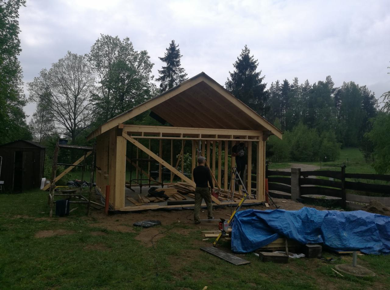 Konstrukcja szkieletowa drewnianego budynku gospodarczego w trakcie montażu, widoczni dwaj pracownicy, jeden na drabinie, drugi na ziemi, w tle las i pochmurne niebo.