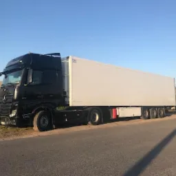 Czarny ciągnik siodłowy Mercedes-Benz z białą naczepą chłodnią zaparkowany na poboczu drogi asfaltowej, w tle zielona bariera dźwiękochłonna i czerwony samochód ciężarowy.