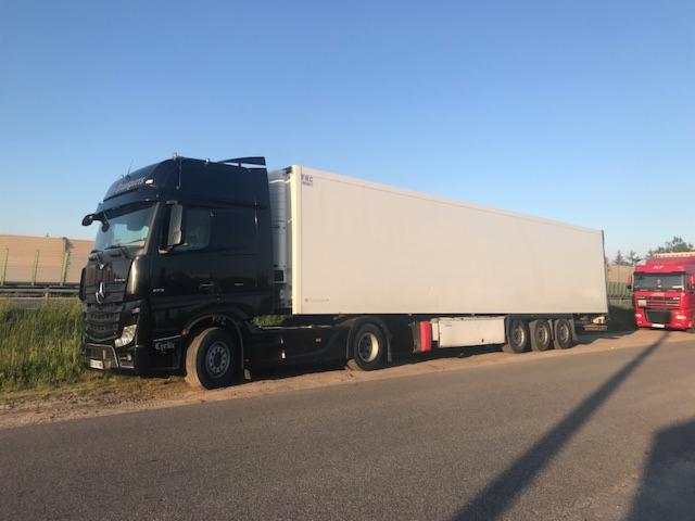 Czarny ciągnik siodłowy Mercedes-Benz z białą naczepą chłodnią zaparkowany na poboczu drogi asfaltowej, w tle zielona bariera dźwiękochłonna i czerwony samochód ciężarowy.
