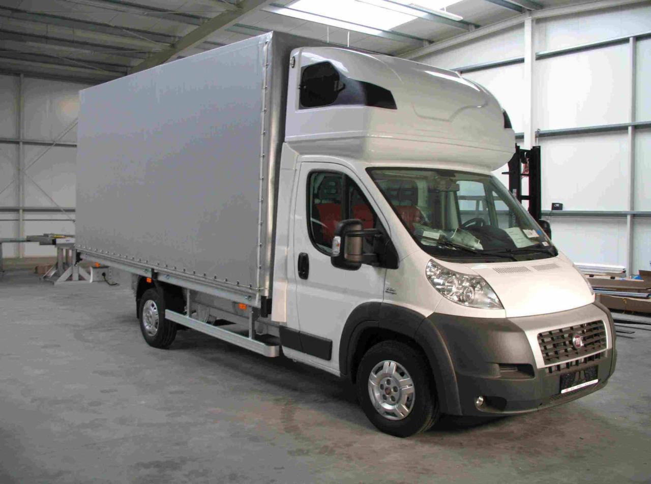 Biały samochód dostawczy z plandeką w hali, widok z przodu z lekkiego ukosa, widoczne elementy konstrukcyjne plandeki.