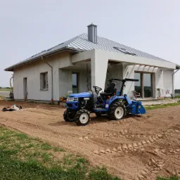 Niebieski mini-traktor z glebogryzarką na przygotowanym podłożu przed nowoczesnym domem z szarą blachodachówką, widoczne ślady opon na piasku i pozostałości trawy.