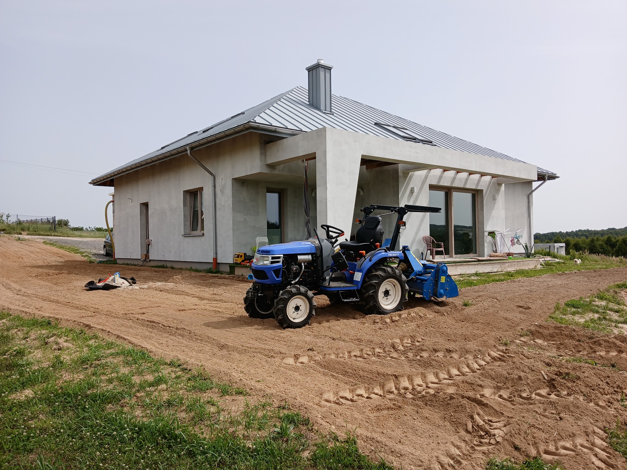Niebieski mini-traktor z glebogryzarką na przygotowanym podłożu przed nowoczesnym domem z szarą blachodachówką, widoczne ślady opon na piasku i pozostałości trawy.