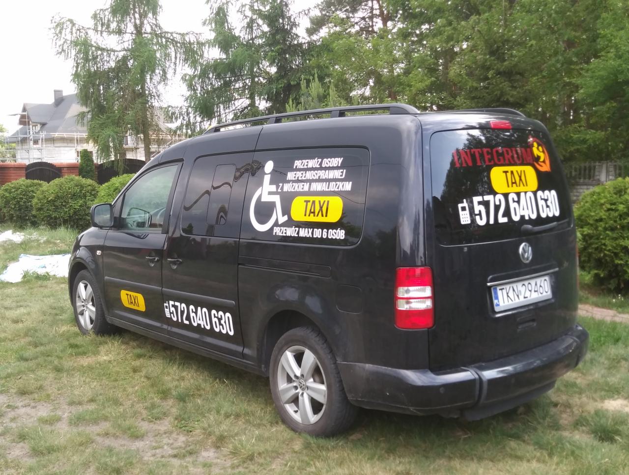 Czarny van z naklejkami informującymi o przewozie osób niepełnosprawnych i numerem telefonu, zaparkowany na trawie przed domem.