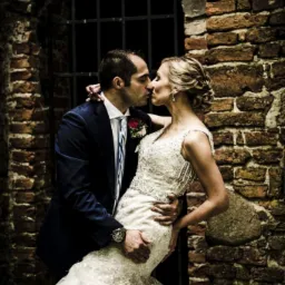 Pocałunek nowożeńców na tle ceglanej ściany z kratą okienną, panna młoda w koronkowej sukni z odkrytymi ramionami, pan młody w granatowym garniturze z zegarkiem na ręku.