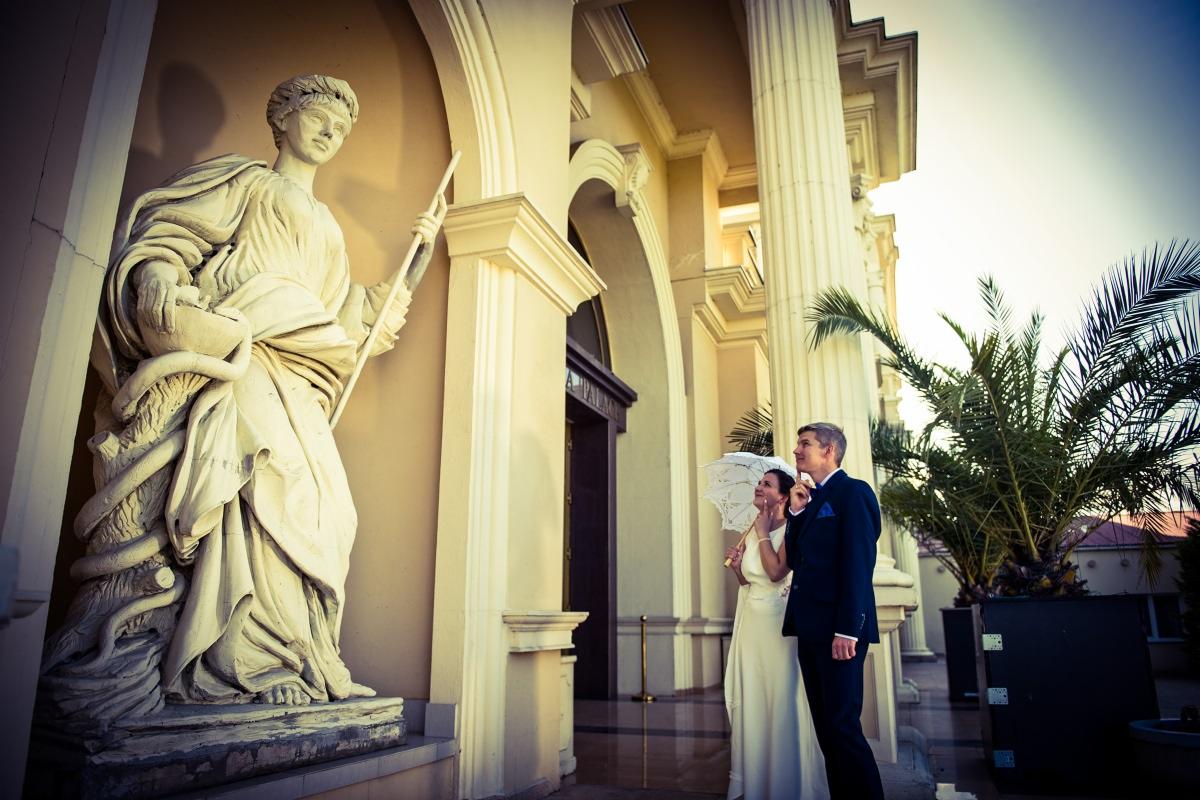 Para młoda z białym parasolem pozuje przy klasycystycznej architekturze z kolumnami i rzeźbą starożytnej postaci.