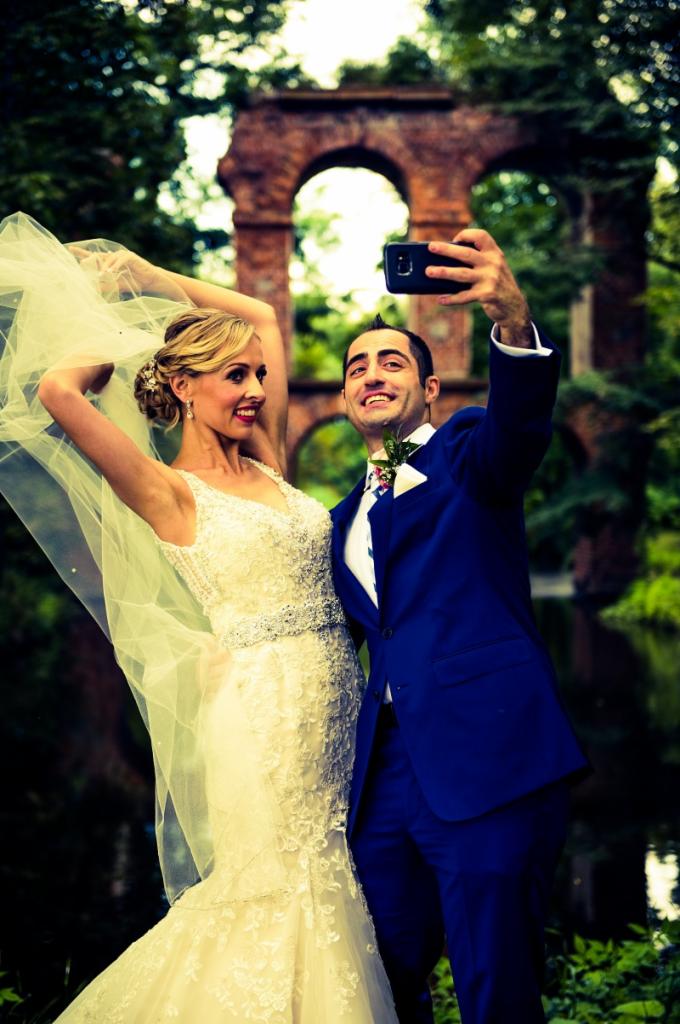 Uśmiechnięta para młoda robi selfie na tle ceglanych ruin w plenerze; panna młoda w koronkowej sukni z welonem, pan młody w granatowym garniturze z butonierką.