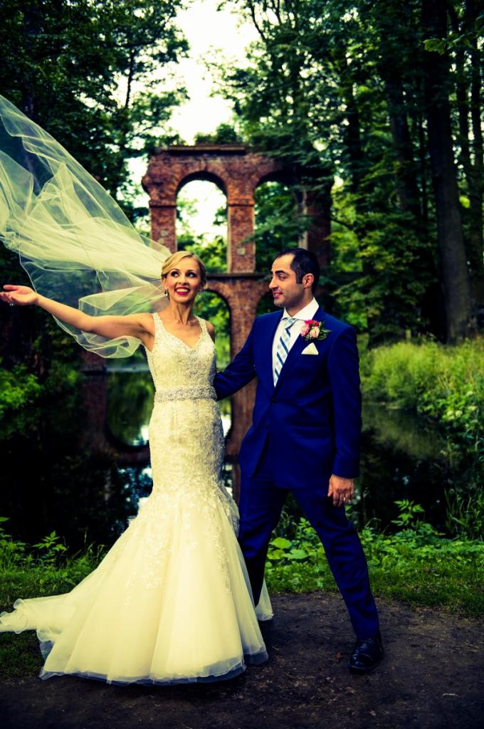 Para młoda w plenerze: uśmiechnięta blondynka w koronkowej sukni z welonem powiewającym na wietrze i pan młody w niebieskim garniturze, na tle ruin ceglanego mostu i zieleni drzew.