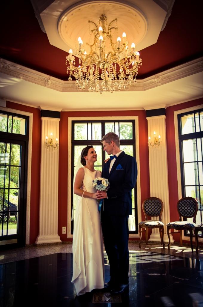 Para młoda w eleganckim wnętrzu z żyrandolem, panna młoda trzyma bukiet z niebieskimi akcentami, oboje patrzą na siebie z uśmiechem.