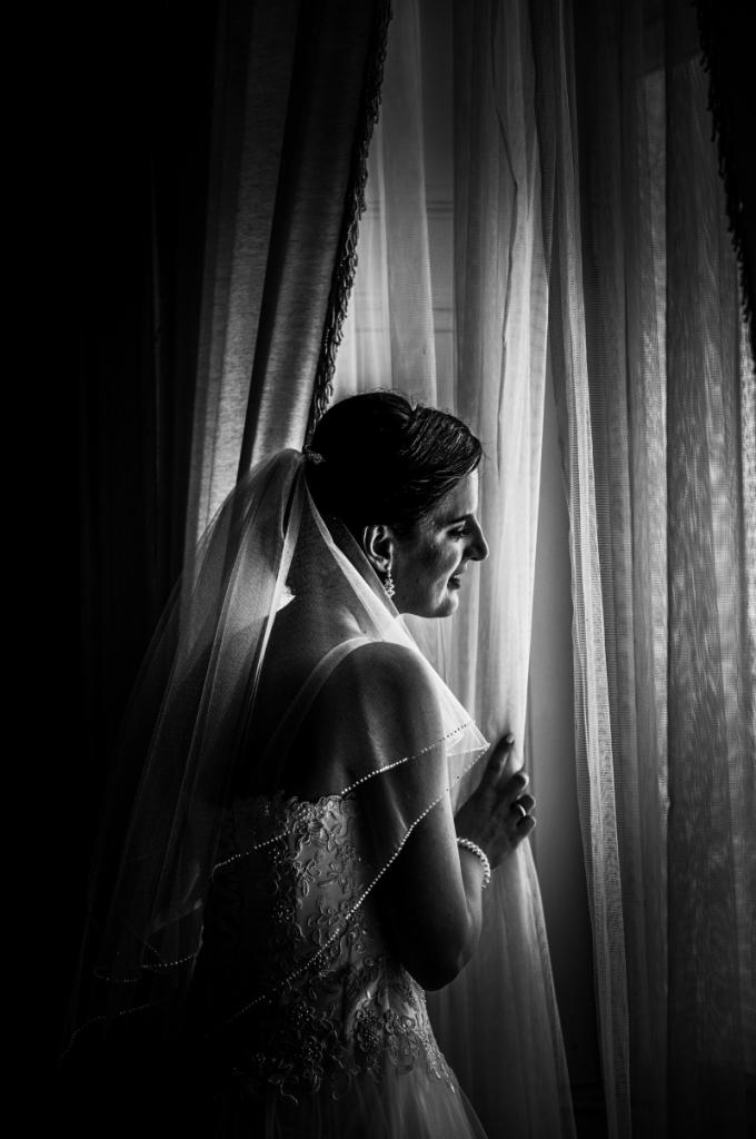 Czarno-białe zdjęcie panny młodej w sukni ślubnej z welonem, stojącej przy oknie z firankami i patrzącej na zewnątrz.