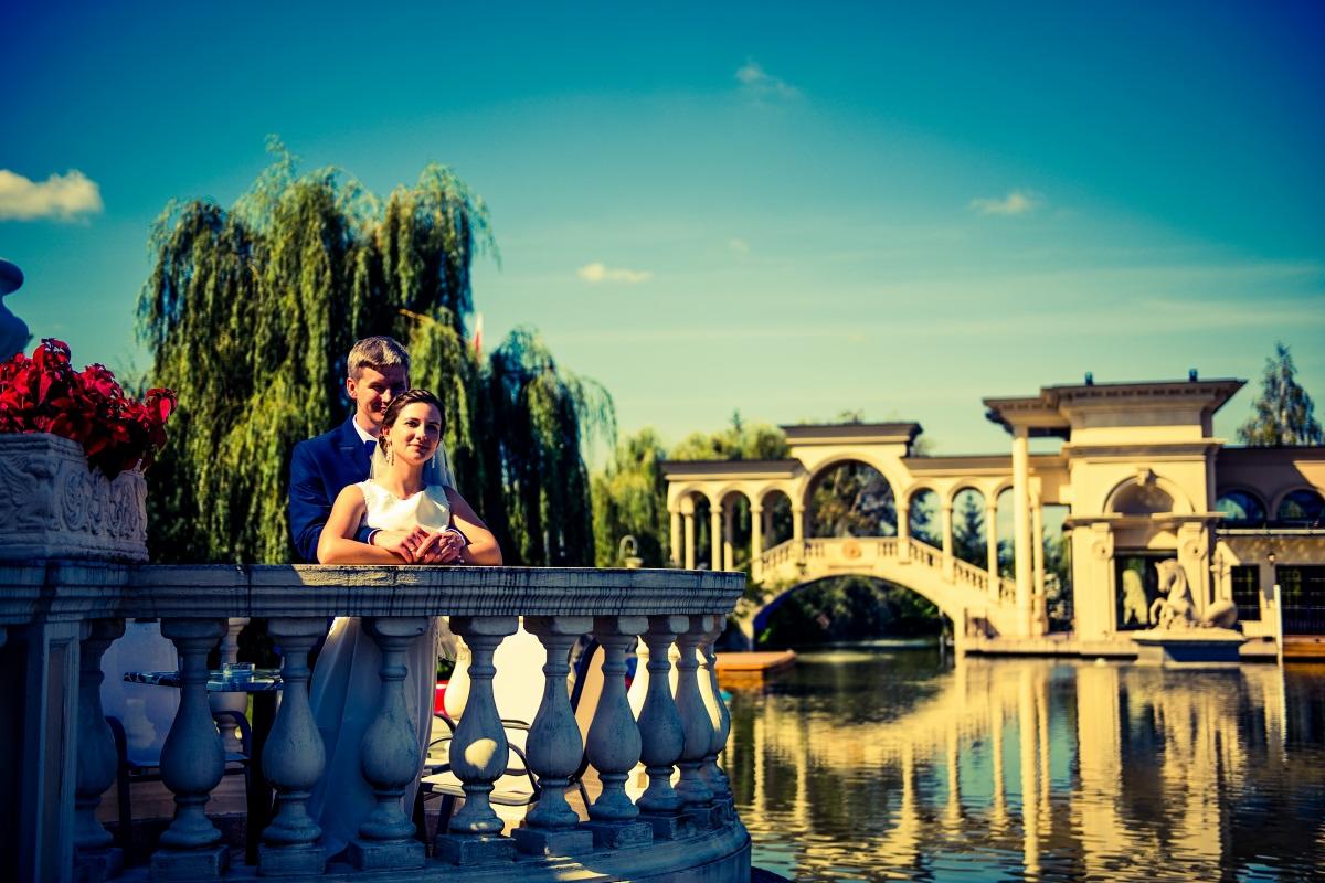 Para młoda w eleganckich strojach pozuje na tle malowniczego parku z wodą i architekturą, mężczyzna w granatowym garniturze obejmuje kobietę w białej sukni, widoczne odbicia w wodzie.