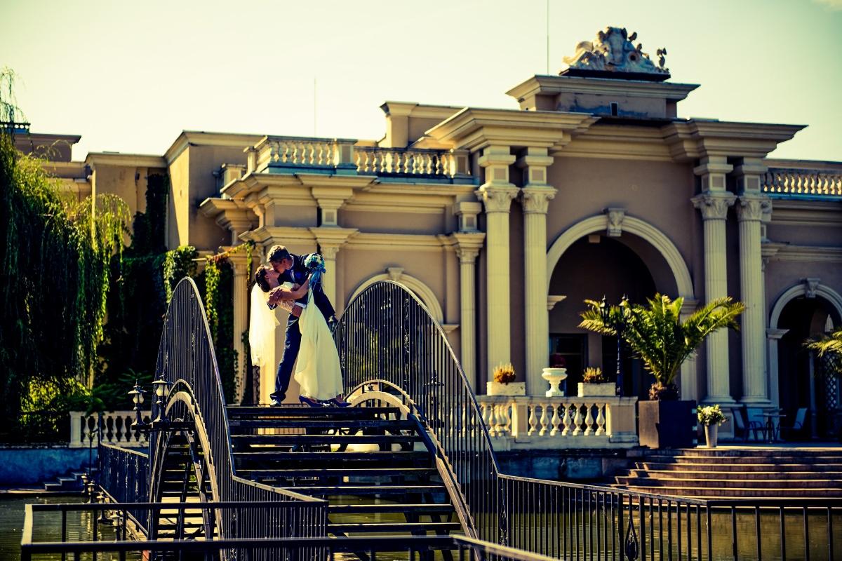 Pocałunek pary młodej na metalowym mostku z widokiem na budynek z kolumnami i roślinnością.