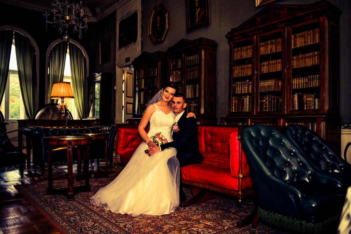 Para młoda siedząca na czerwonej sofie w bogato zdobionym wnętrzu biblioteki z regałami pełnymi książek, pod żyrandolem. Panna młoda w białej sukni z bukietem, pan młody w ciemnym garniturze.