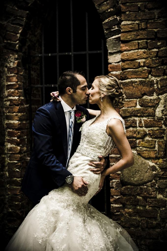 Pocałunek nowożeńców na tle ceglanej ściany z kratą okienną, panna młoda w koronkowej sukni z odkrytymi ramionami, pan młody w granatowym garniturze z zegarkiem na ręku.