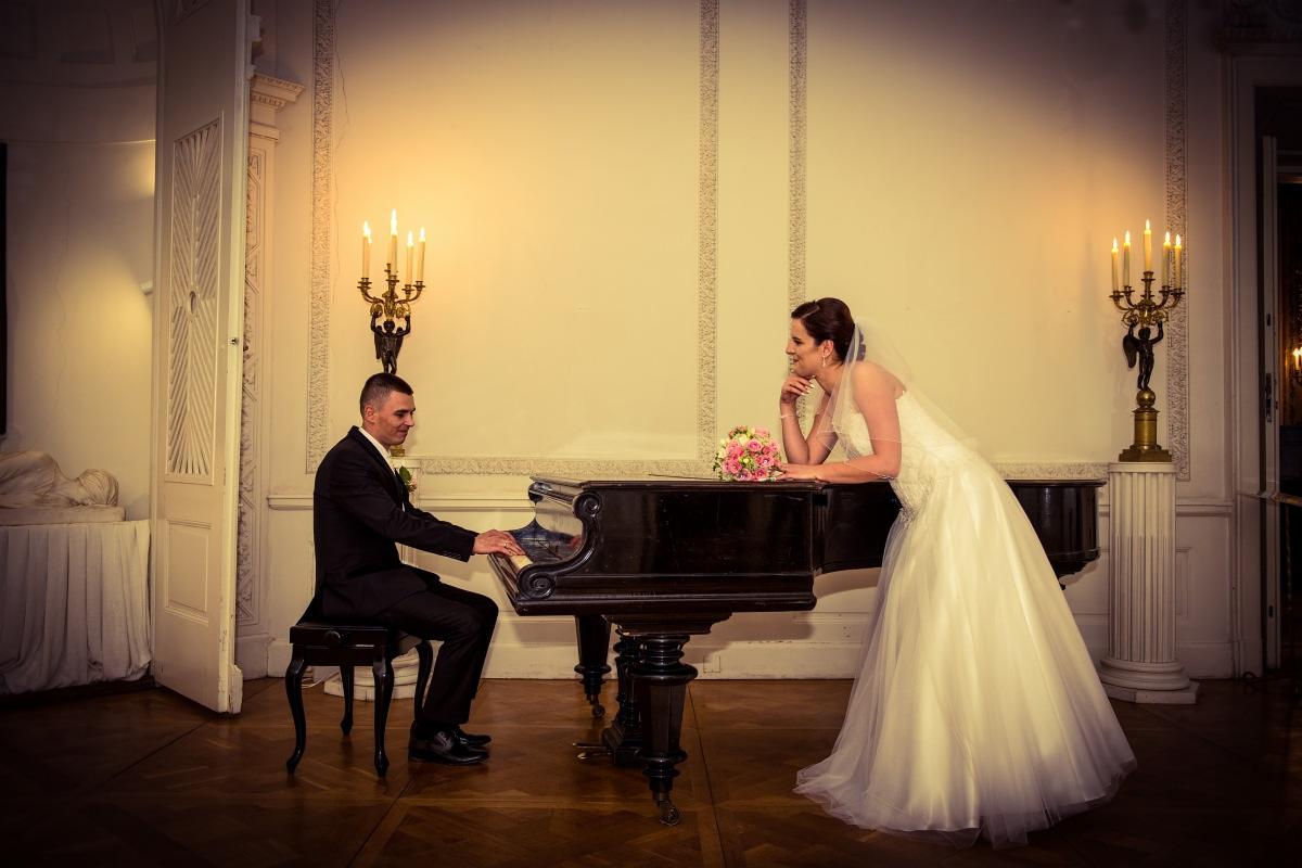Panna młoda w białej sukni opiera się o czarny fortepian, na którym gra pan młody w ciemnym garniturze, w tle świeczniki i klasyczne wnętrze.