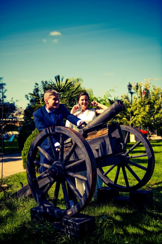 Para młoda pozuje przy starej armacie na trawniku w słoneczny dzień; pan młody w niebieskim garniturze, panna młoda w białej sukni.