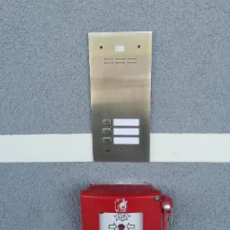 Zewnętrzny panel domofonowy ze stali nierdzewnej, zamontowany nad czerwoną skrzynką alarmową przeciwpożarową na szarej, teksturowanej ścianie budynku.
