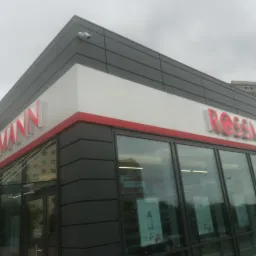 Kamera monitoringu zamontowana na elewacji budynku z logo Rossmann, widoczne okna wystawowe i szara elewacja.