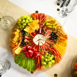 Dekoracyjna drewniana deska z serami, wędlinami, owocami, papryką i krakersami, ułożona na stole z konfetti i kieliszkami do wina, widok z g&oacute;ry.