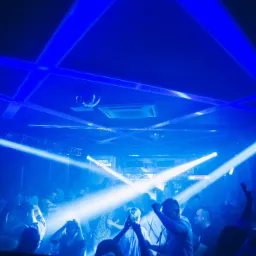 Tłum ludzi w klubie nocnym oświetlonym intensywnym niebieskim światłem, z reflektorami przecinającymi przestrzeń i dyskotekową kulą na suficie.