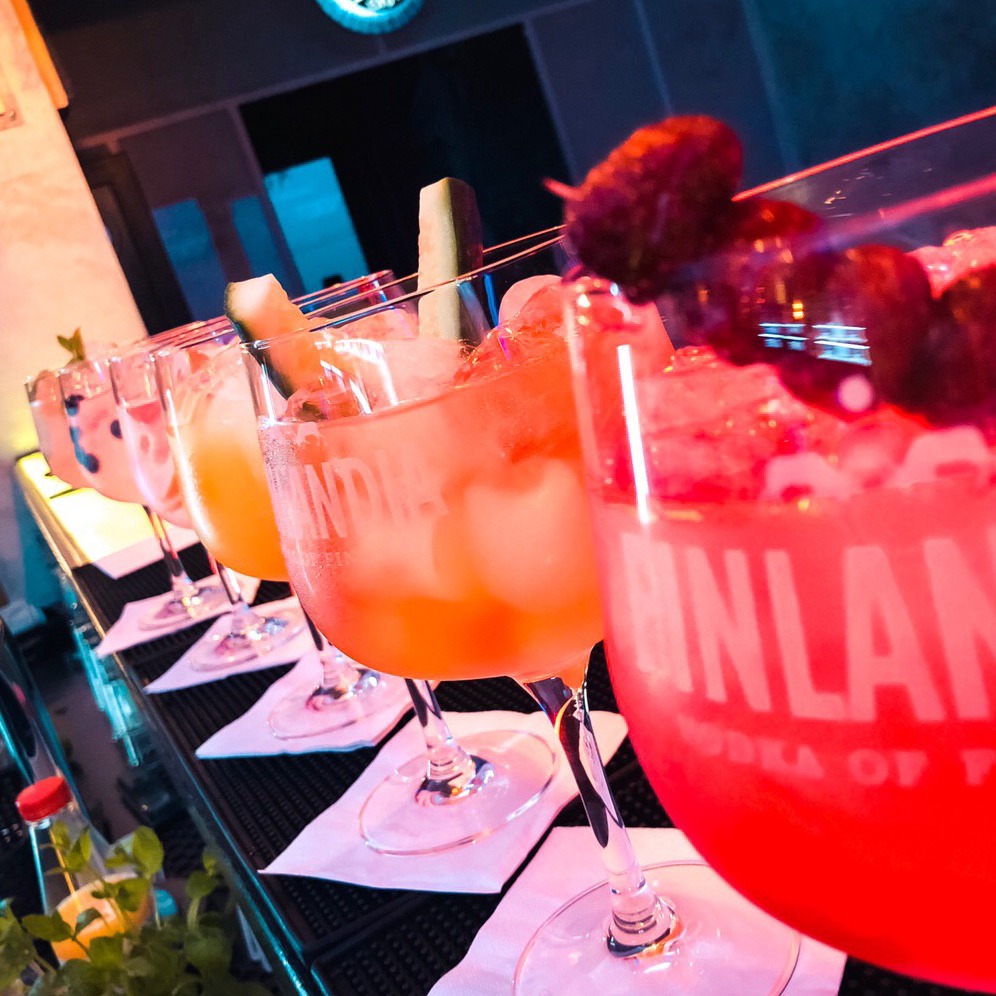 Rząd kolorowych koktajli w kieliszkach na nóżce, udekorowanych owocami i kostkami lodu, stojących na barze podświetlonym neonowym światłem.