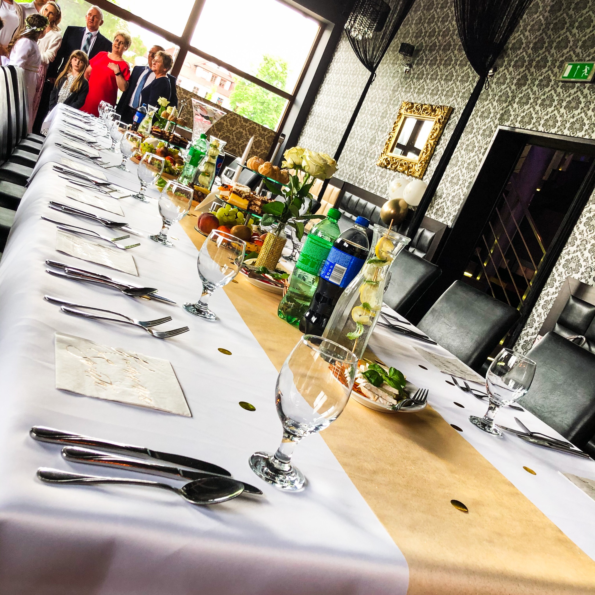 Uroczysty stół z białym obrusem, złotym bieżnikiem, nakryciami, szklankami, owocami, ciastami i napojami, z grupą osób w tle przy oknie.