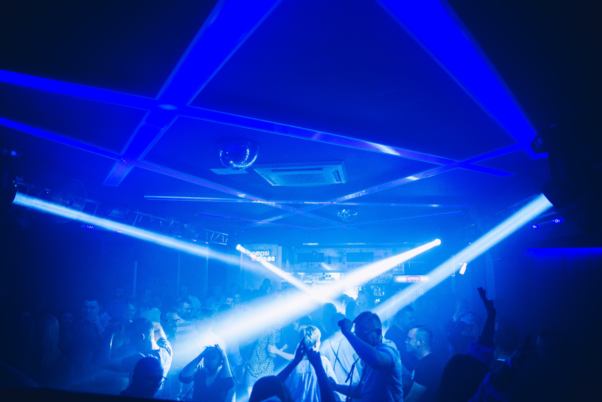 Tłum ludzi w klubie nocnym oświetlonym intensywnym niebieskim światłem, z reflektorami przecinającymi przestrzeń i dyskotekową kulą na suficie.