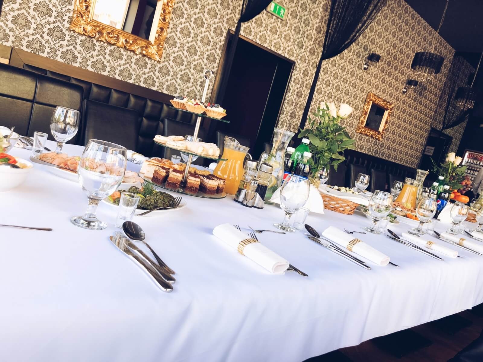 Uroczysty stół z białym obrusem, nakryty zastawą, przekąskami, ciastami i napojami, dekorowany kwiatami, w tle wzorzysta tapeta i lustra w złotych ramach.