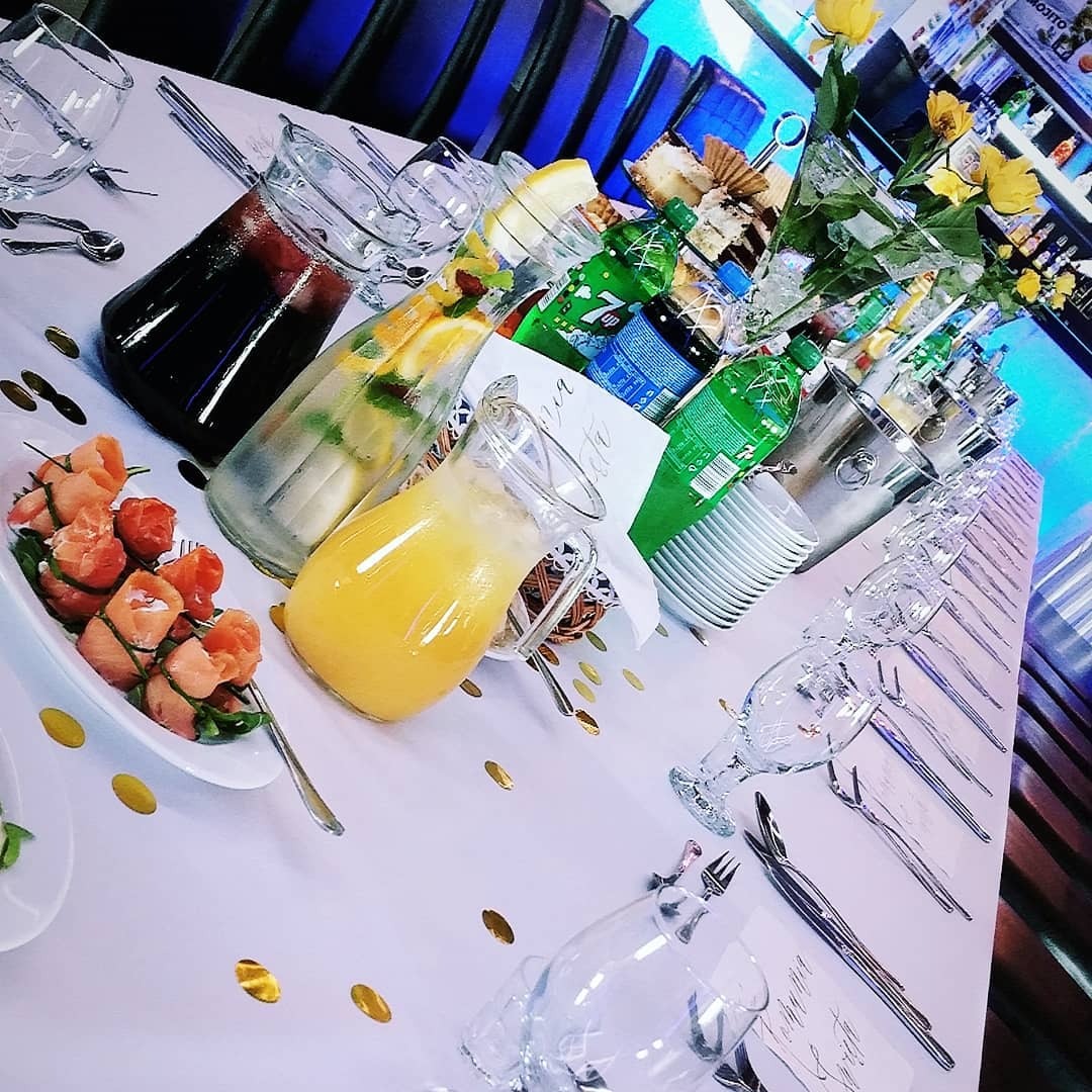 Uroczysty stół z napojami w dzbankach, przekąskami z łososia, butelkami w wiaderku z lodem i żółtymi różami w wazonie, udekorowany złotymi konfetti.