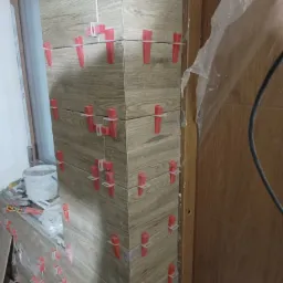 Układanie płytek imitujących drewno na ścianie, z widocznymi czerwonymi klinami poziomującymi i białymi krzyżykami dystansowymi, obok okna i drewnianej futryny drzwi.