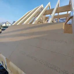 Konstrukcja drewniana dachu w trakcie budowy, pokryta płytami izolacyjnymi z nadrukiem producenta, widoczne elementy więźby dachowej i fragment krajobrazu z zabudowaniami.